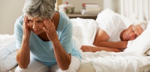 Worried Senior Woman Sits On Bed Whilst Husband Sleeps